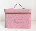 A pink faux ostrich leather cosmetic case with a top handle, gold zipper, and a blank rectangular patch on the front, set against a plain white background.