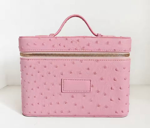A pink faux ostrich leather cosmetic case with a top handle, gold zipper, and a blank rectangular patch on the front, set against a plain white background.