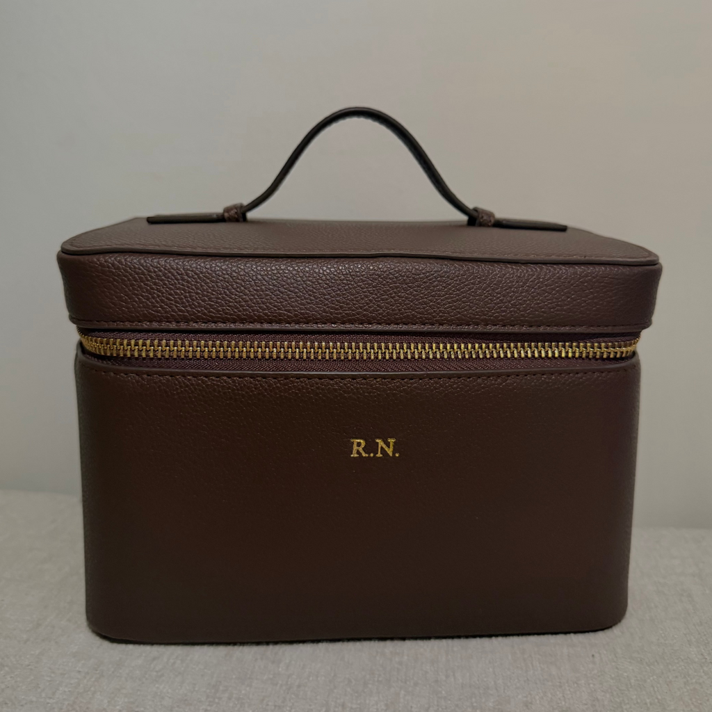 A dark brown textured leather cosmetic case with a top handle, gold zipper, and 'R.N.