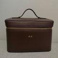A dark brown textured leather cosmetic case with a top handle, gold zipper, and 'R.N.