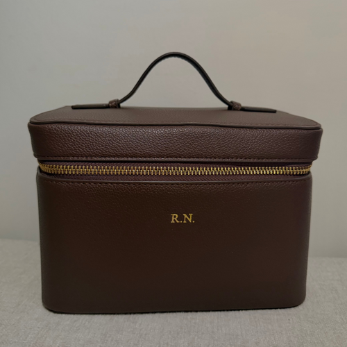 A dark brown textured leather cosmetic case with a top handle, gold zipper, and 'R.N.